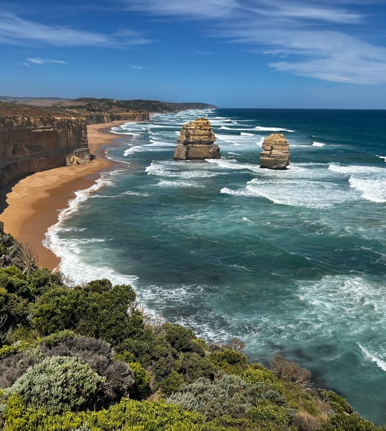 Looking east along the coast. From left  to right. A high bluff above a sandy beach, then two large flowerpot shaped columns in the blue water.