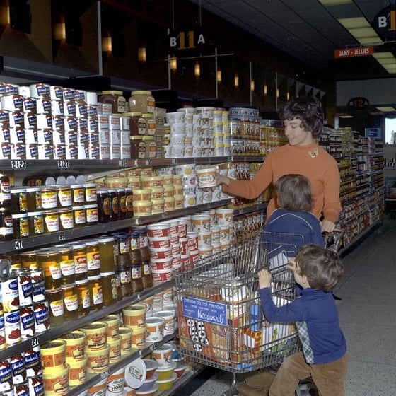 A woman with two children and a shopping cart at a supermarket shelf stacked with tubs and jars of honey.