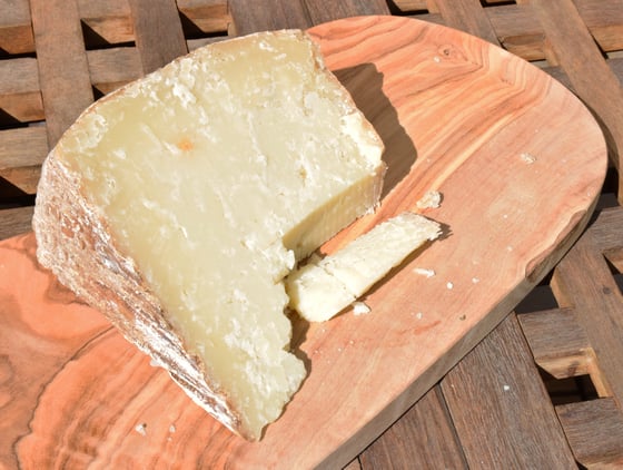 A wedge of pale aged sheep's cheese with a natural rind and one thin slice cut off, on an olive-wood board.