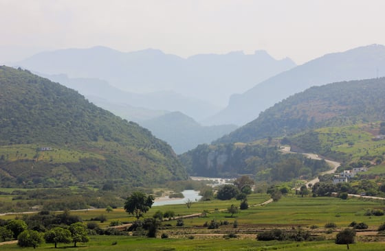 A hazy river valley with green fields in the foreground and forested hills rising to limestone peaks behind.