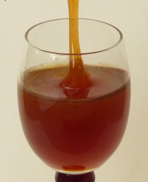 Dark reddish-amber honey pouring in a thick stream into a clear stemmed glass two-thirds full.