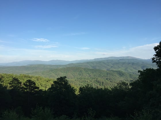 Layered forested ridges under a blue sky, a hazy mountain ridge on the horizon.