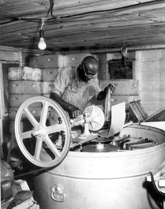 Black-and-white photograph of a man working a hand-cranked honey extractor in a plank-walled room stacked with hive boxes.