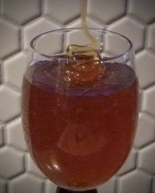 A thin stream of honey falling into a stemmed glass filled with dark reddish honey, against white hexagonal wall tiles.