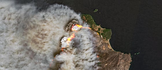 From above, a large white plume drifts over dark sea from a green and brown headland, with lines of orange marking the fire front.