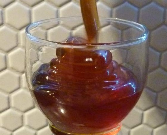 A thick dark stream of honey falling onto a coated dipper standing in a small glass tumbler, against white hexagonal tiles.