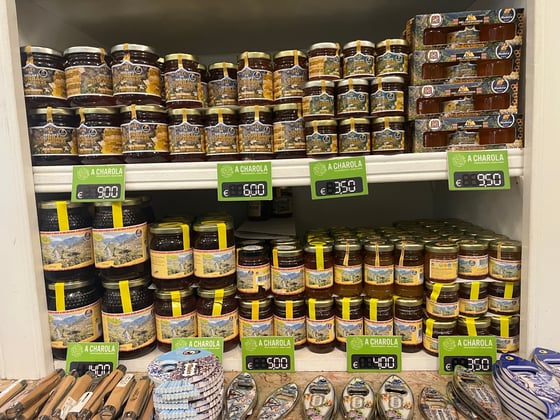 Two shop shelves packed with jars of honey, dark to golden, the upper with blue-and-yellow Curral das Freiras labels and boxed sets at the right, the lower with yellow-banded jars of several sizes; green price cards clipped to each shelf edge and knives and bottle openers on the counter beneath.