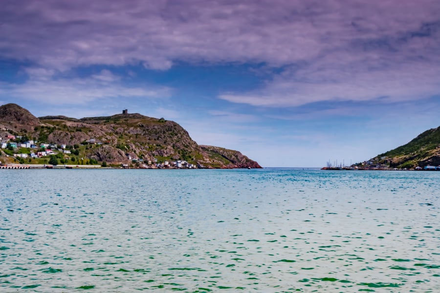 Rocky harbour entrance with a stone tower on the hill above and small houses along the shore.