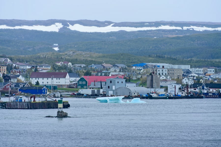 A small iceberg floating in a harbour in front of wharves, fishing boats and town buildings, with snow patches on the hills behind.