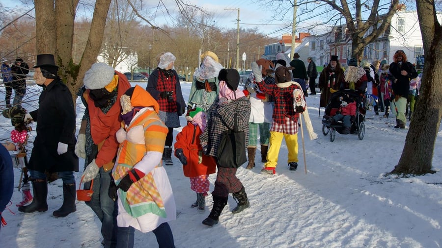 Costumed people with covered faces walking through snow among bare trees, with onlookers behind.