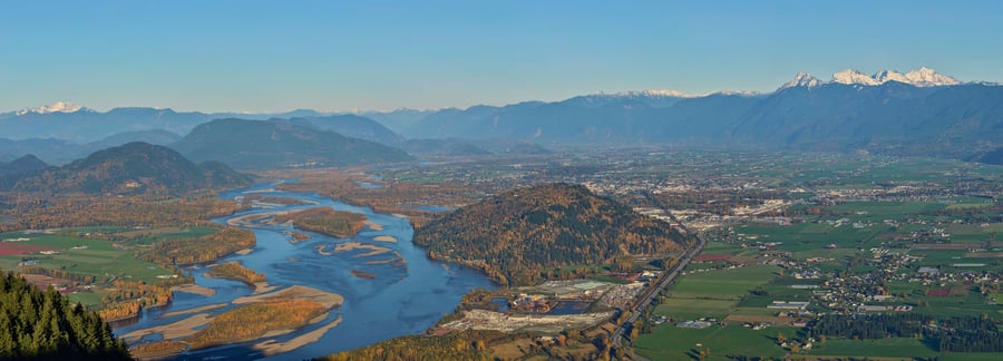 A wide view over the braided Fraser River, farmland on both banks, snow-capped peaks along the horizon.