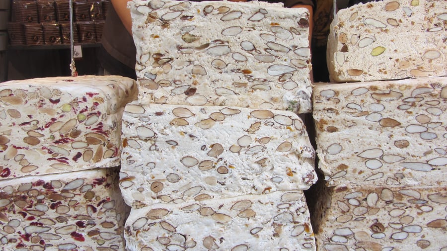 Stacked slabs of white nougat studded with whole almonds, some with green pistachios and red fruit pieces, on a shop counter.