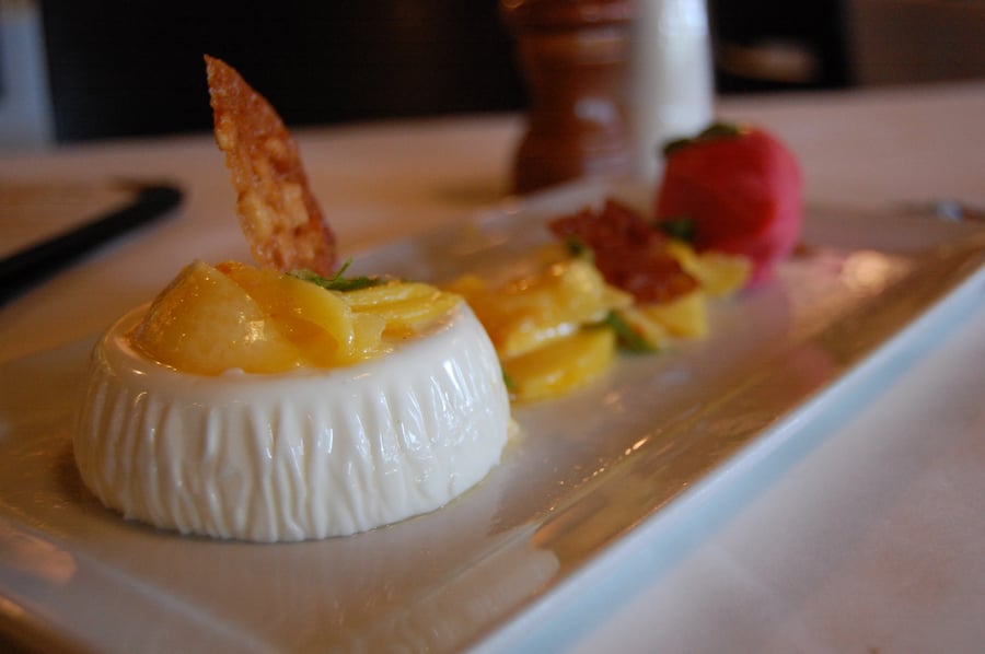 A white fluted panna cotta topped with diced yellow fruit and a thin crisp, beside a fruit salad and a scoop of red sorbet on a long white plate.