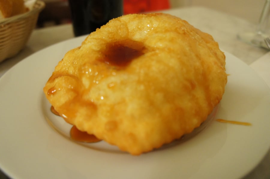 A puffed, golden fried pastry on a white plate with dark amber honey pooled on top and running onto the plate.