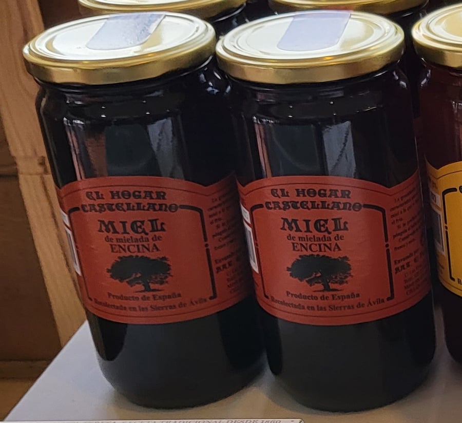 Two tall glass jars of near-black honey with gold lids, standing side by side on a wooden table.