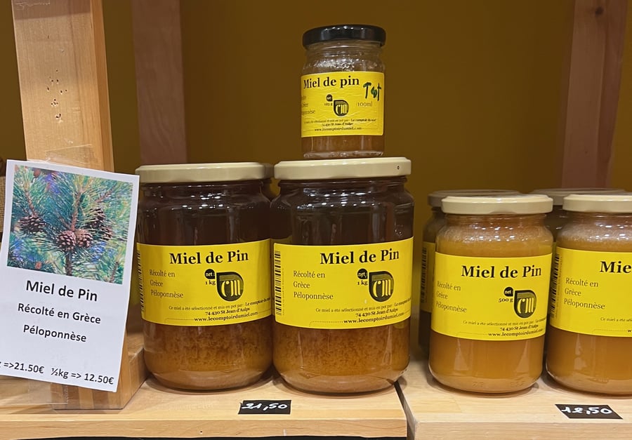 Three jars of amber-brown honey with yellow labels on a wooden shelf, beside a card with a pine-cone picture and chalked prices.