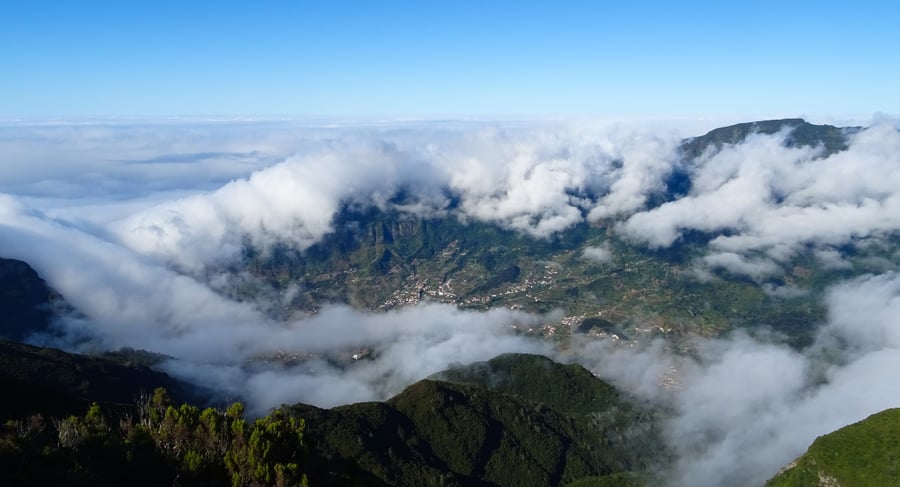 A wide view down into a green valley from a mountain rim, white cloud over the ridges and lying in the valley, villages and terraces visible between the clouds, shrubs in the foreground and blue sky above.